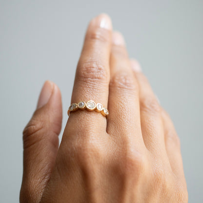 A close-up of a hand wearing a delicate gold band ring adorned with small round clear stones, set against a soft gray background.
