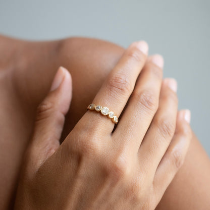 A delicate gold ring with three small round-cut clear gemstones worn on a person's finger, shown in a close-up lifestyle shot against a soft gray background.