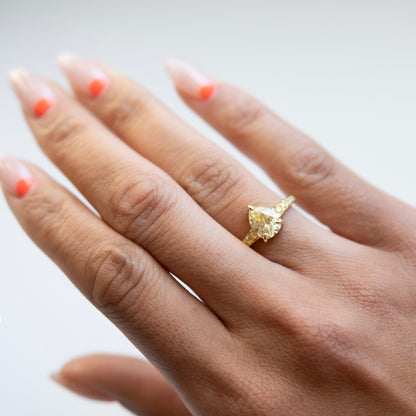 Gold ring with a pear-shaped diamond on a hand with orange nail polish against a light background
