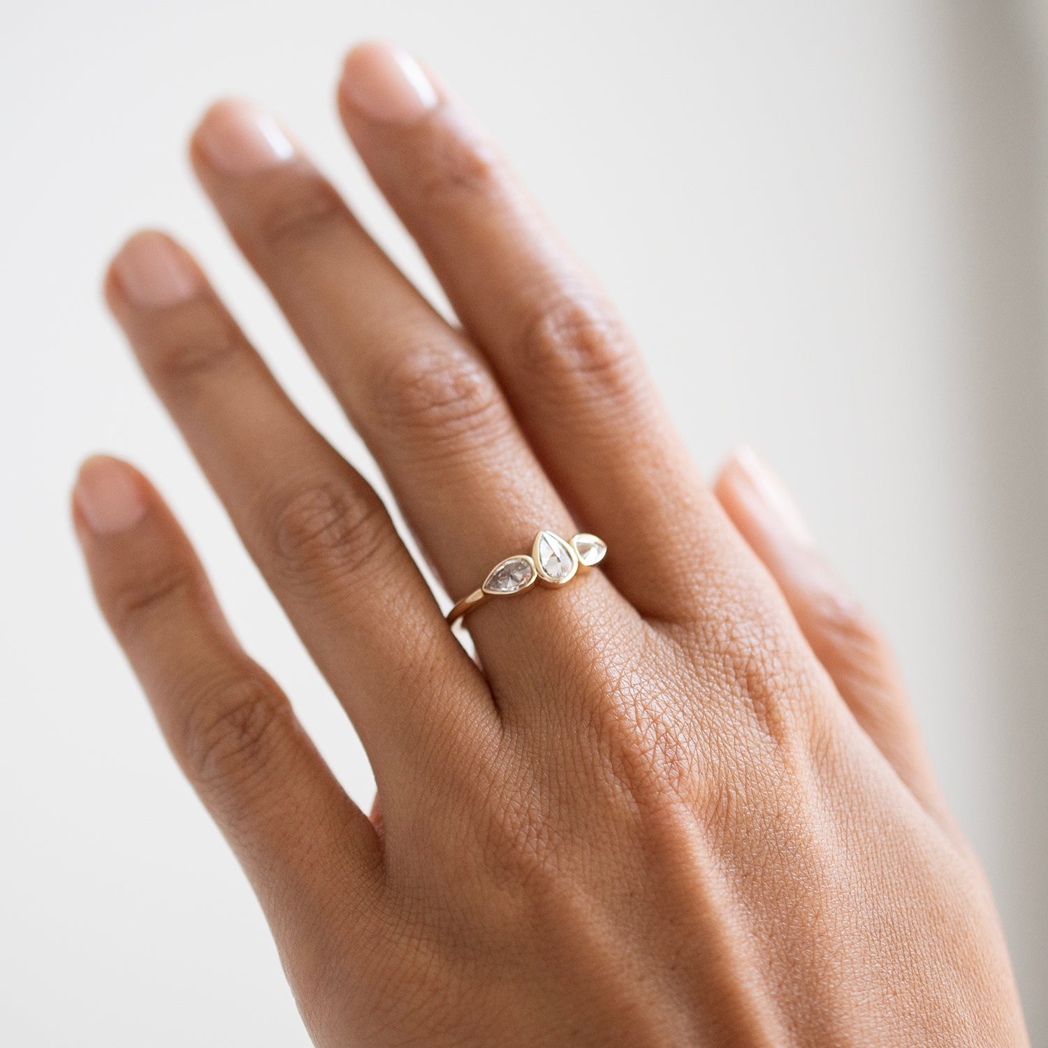 A close-up of a hand wearing a delicate gold ring with three small clear marquise-cut gemstones on a soft light gray background.