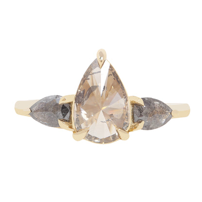 A gold ring featuring a large pear-cut clear diamond with inclusions, accented by two smaller marquise-cut gray diamonds on either side, shown on a plain white background.