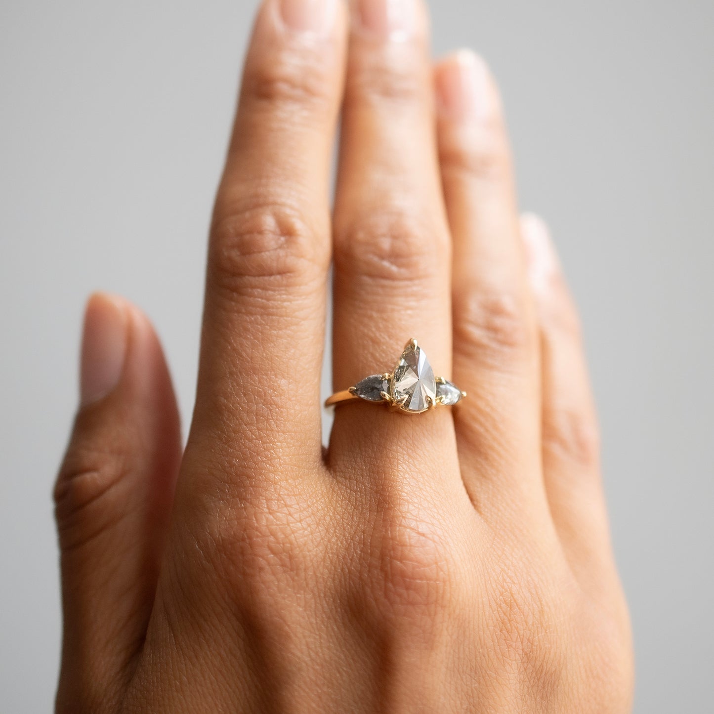 A close-up of a hand wearing a delicate gold ring with three diamonds clustered near the base of the finger, set against a soft gray background.