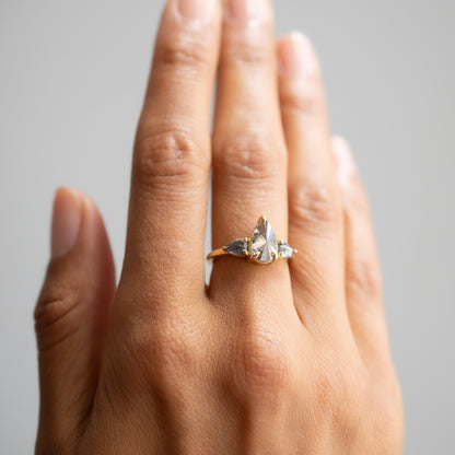 A close-up of a hand wearing a delicate gold ring with three diamonds clustered near the base of the finger, set against a soft gray background.