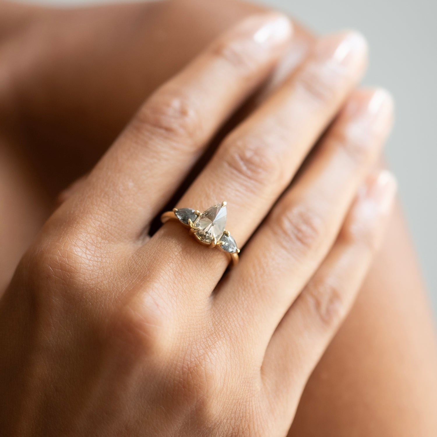 A close-up of a hand wearing a delicate gold ring with a central diamond and small diamonds, photographed against a soft neutral background.