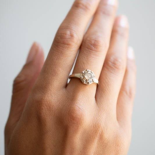 A close-up of a hand wearing a white gold ring with a round clear gemstone, set against a soft light gray background.