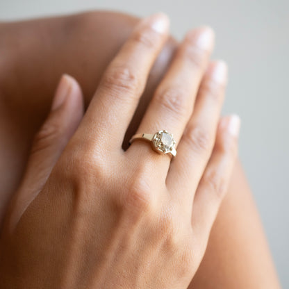 A white gold ring with a round clear gemstone set on a person's ring finger, shown in a close-up lifestyle shot against a soft neutral background.