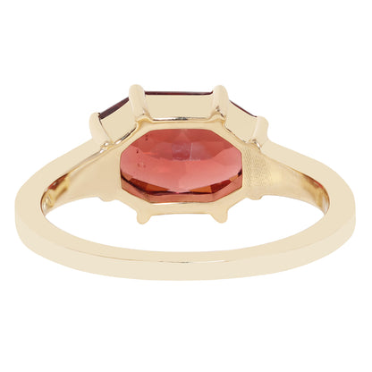 A gold ring featuring a large, faceted red central gemstone in a prong setting shown against a plain white background.