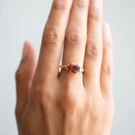 A close-up of a hand wearing a gold ring with a marquise-cut gemstone that has a red center set against a soft light gray background.
