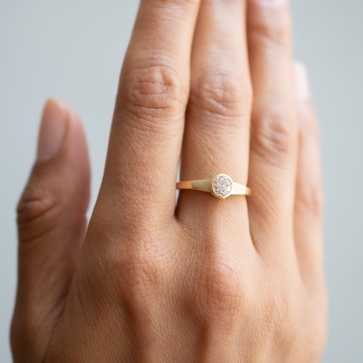 A close-up of a hand wearing a delicate gold ring with a round clear diamond, set against a soft light gray background.