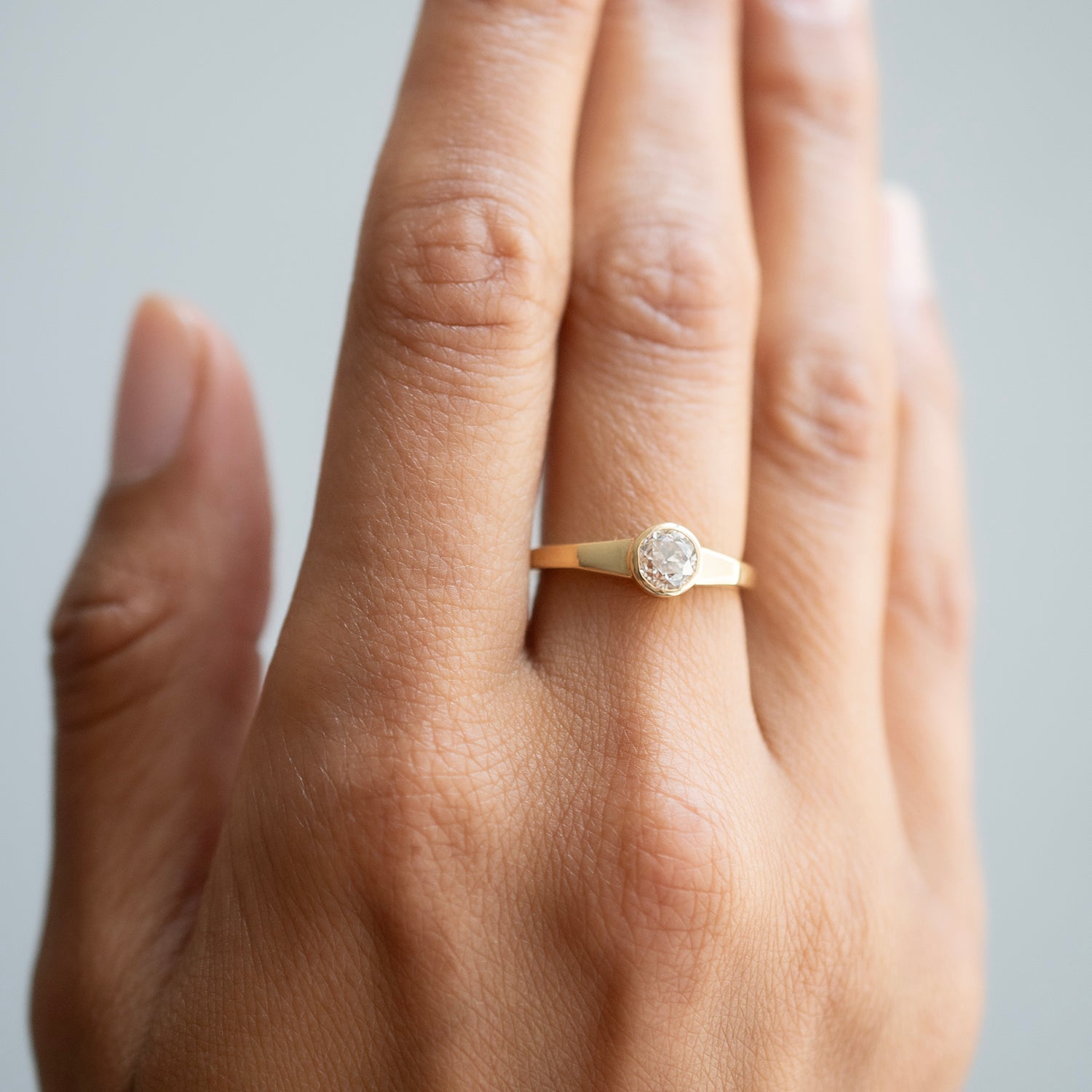 A close-up of a hand wearing a delicate gold ring with a round clear diamond, set against a soft light gray background.