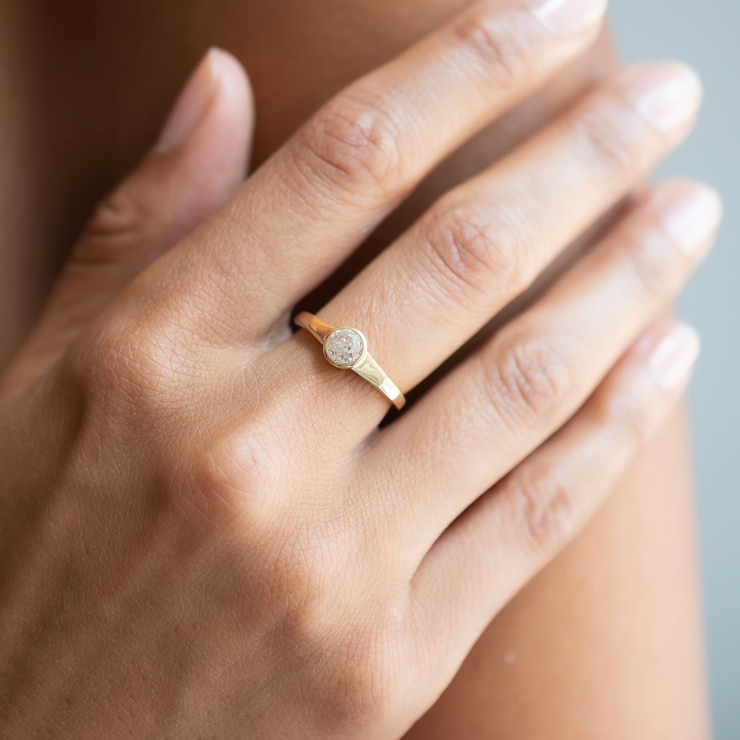 A close-up of a hand wearing a delicate gold ring with a round bezel-set clear diamond.