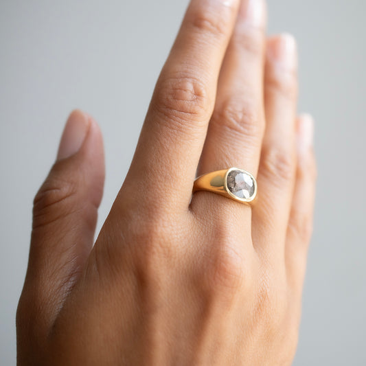 A close-up of a hand wearing a gold signet ring with a round, bezel-set clear gemstone, set against a soft, light gray background.