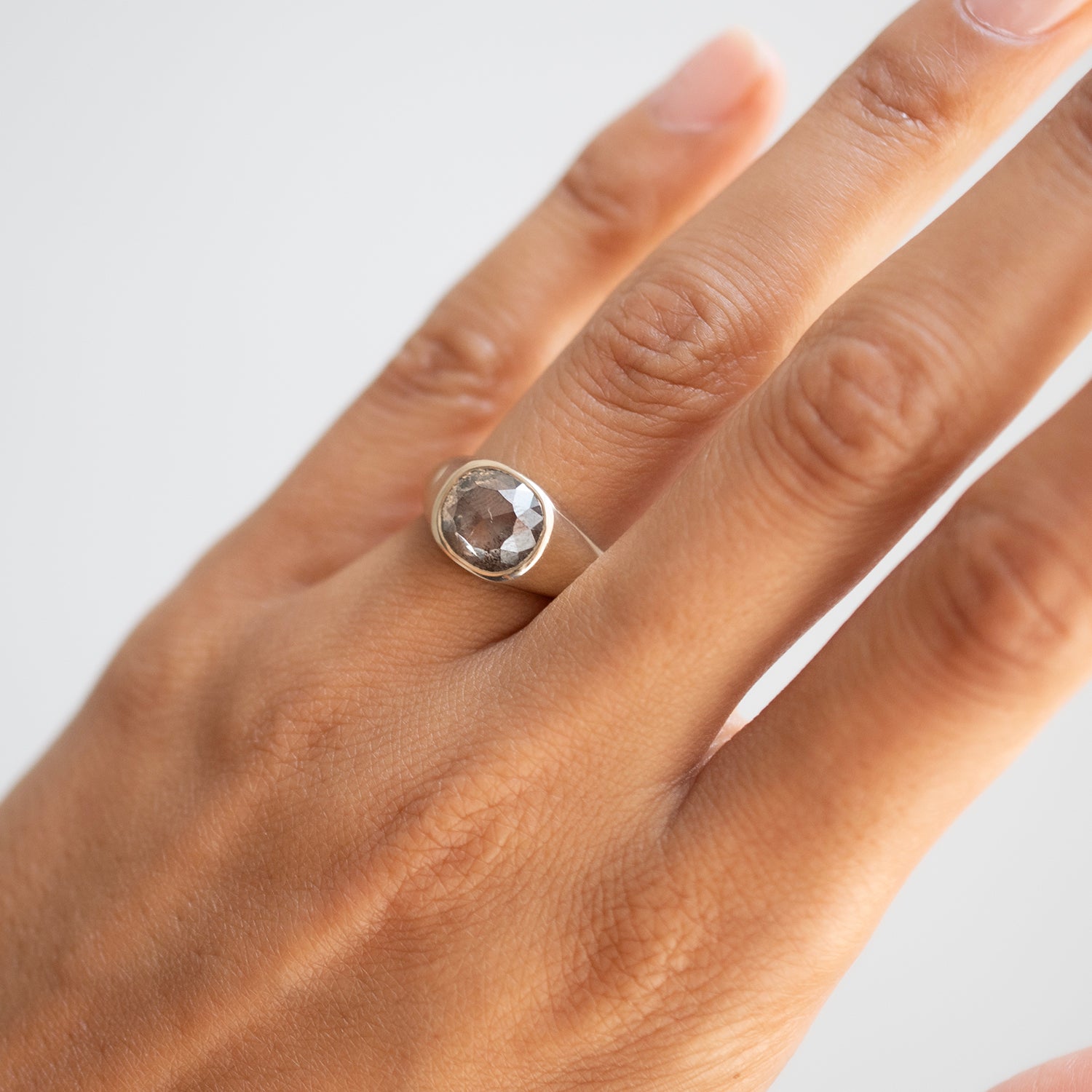 A silver ring with a round faceted clear gemstone set on a person's ring finger, shown against a plain light gray background.