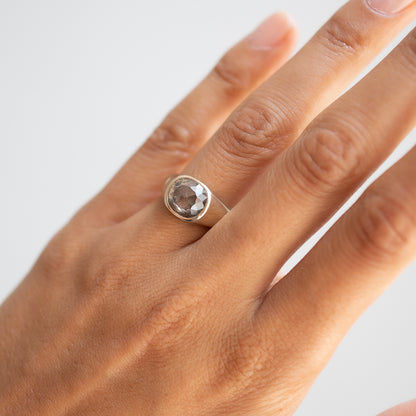 A silver ring with a round faceted clear gemstone set on a person's ring finger, shown against a plain light gray background.