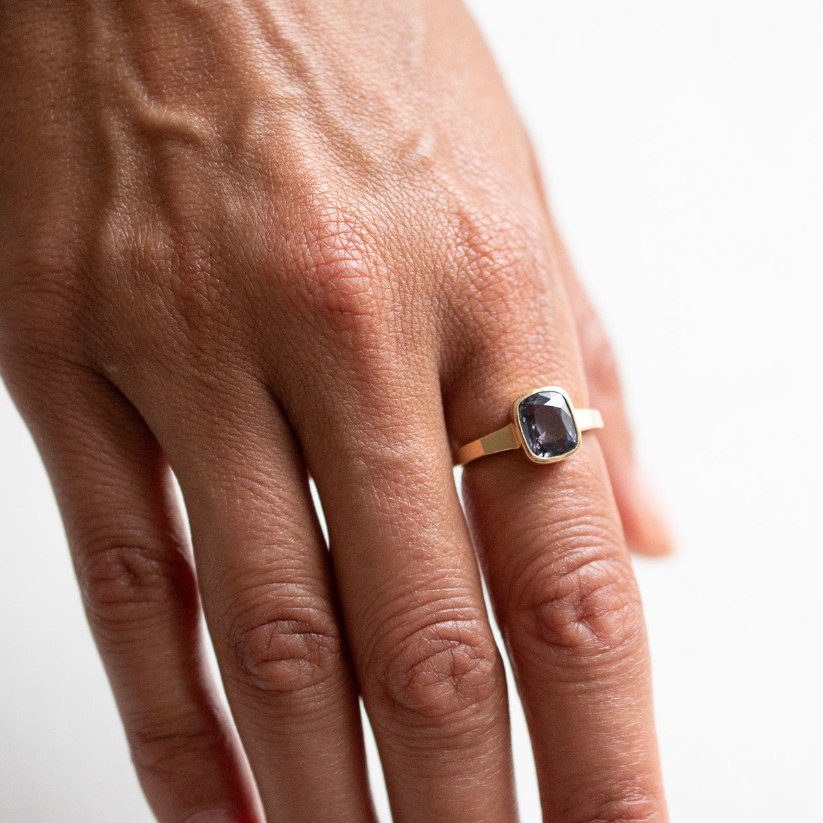 A close-up of a person's hand wearing a gold ring with a rectangular dark blue stone on a plain white background.