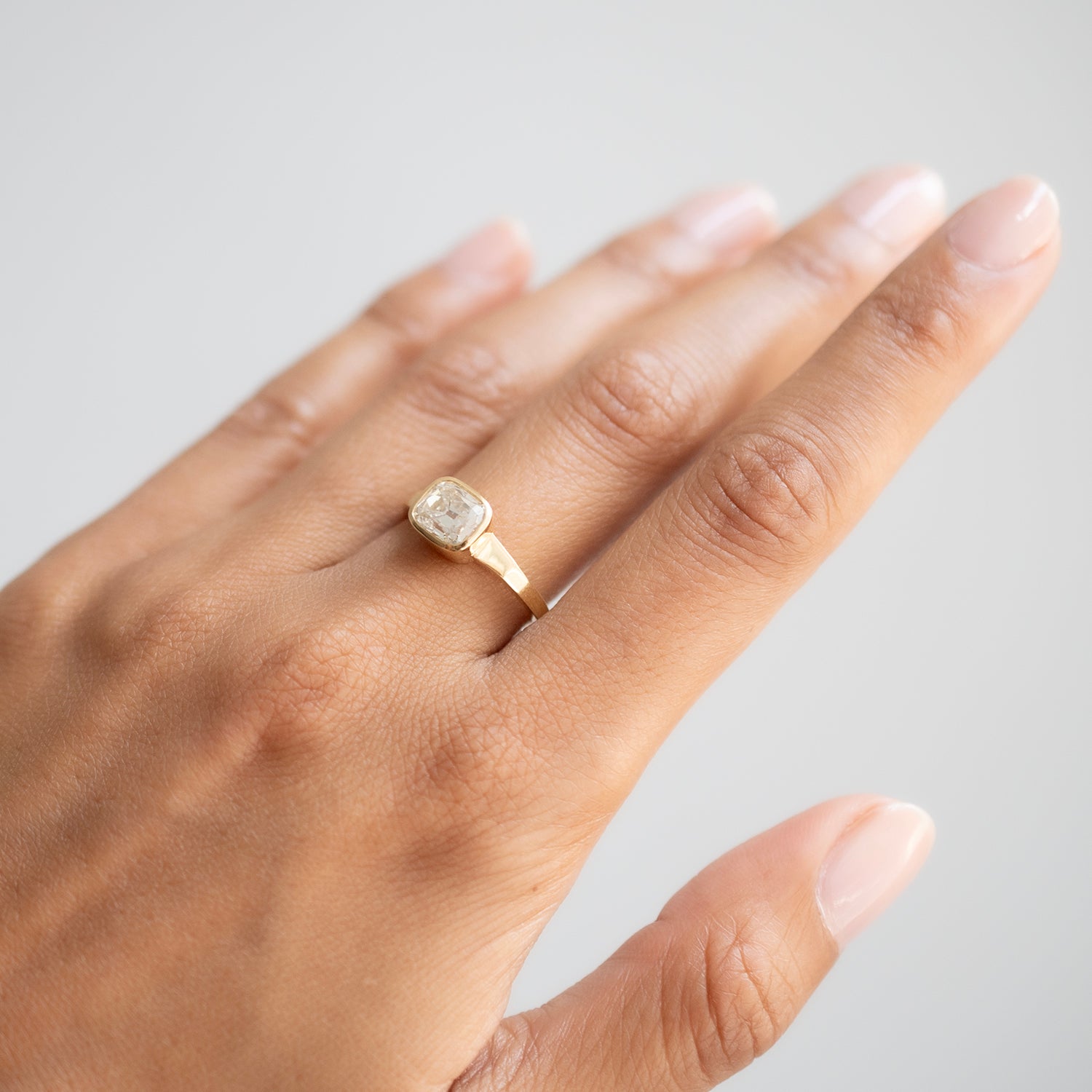 A gold ring with a square bezel-set clear gemstone worn on a person's ring finger, shown against a neutral light gray background.