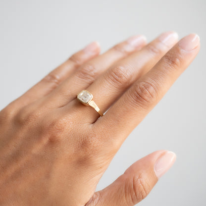 A gold ring with a square bezel-set clear gemstone worn on a person's ring finger, shown against a neutral light gray background.