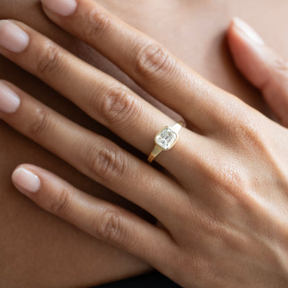 A close-up of a hand wearing a gold ring with a rectangular clear gemstone, resting on a soft beige surface.