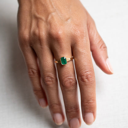 A close-up of a hand wearing a gold ring with a rectangular green gemstone, set against a plain light background.