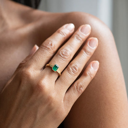 A close-up of a hand wearing a gold ring with a rectangular green emerald-cut gemstone, resting on the person's shoulder.
