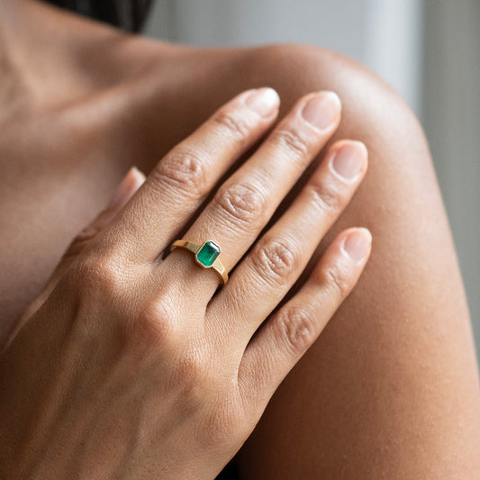 A close-up of a hand wearing a gold ring with a rectangular green emerald-cut gemstone, resting on the person's shoulder.