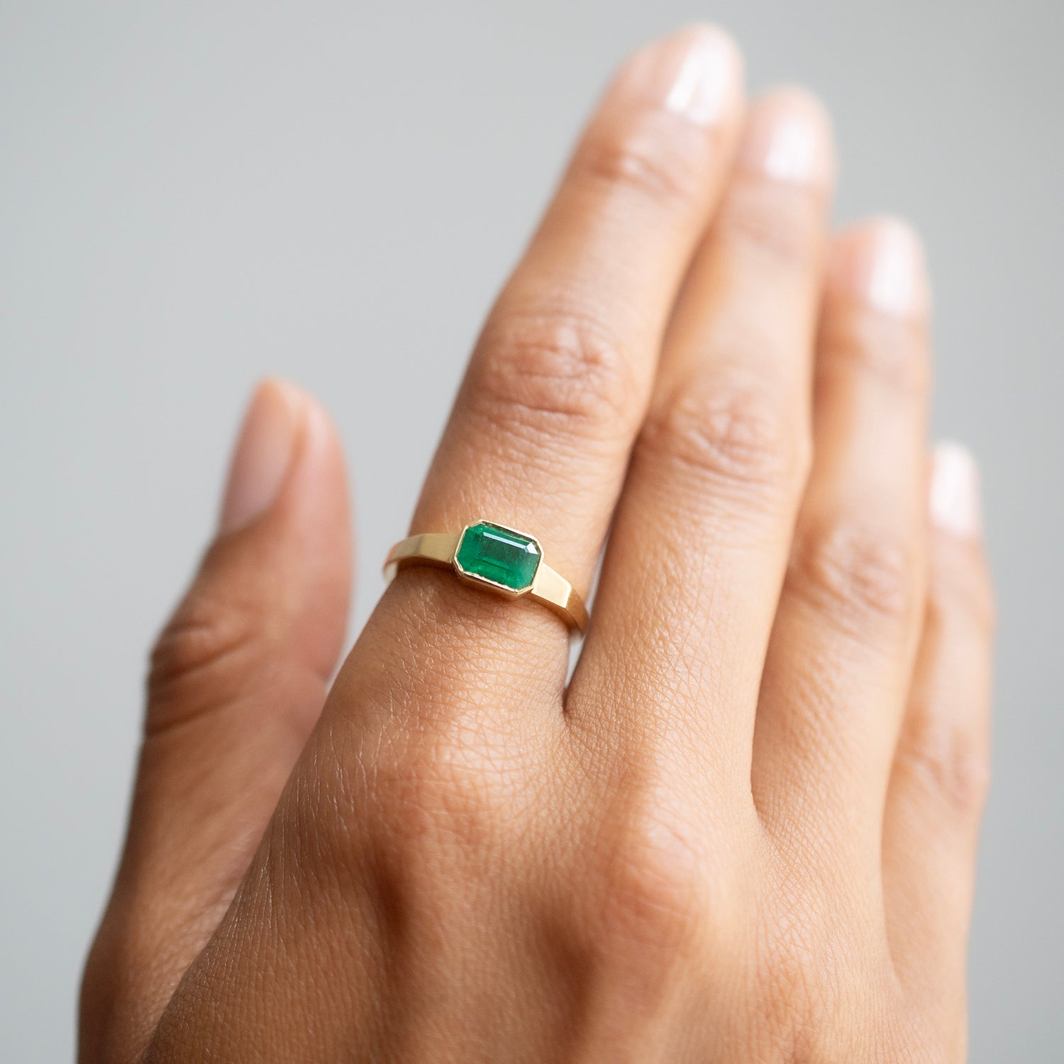 A gold ring with a rectangular green emerald-cut gemstone, shown worn on a person's ring finger against a soft gray background.