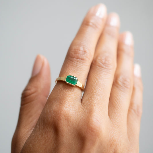 A gold ring with a rectangular green emerald-cut gemstone, shown worn on a person's ring finger against a soft gray background.