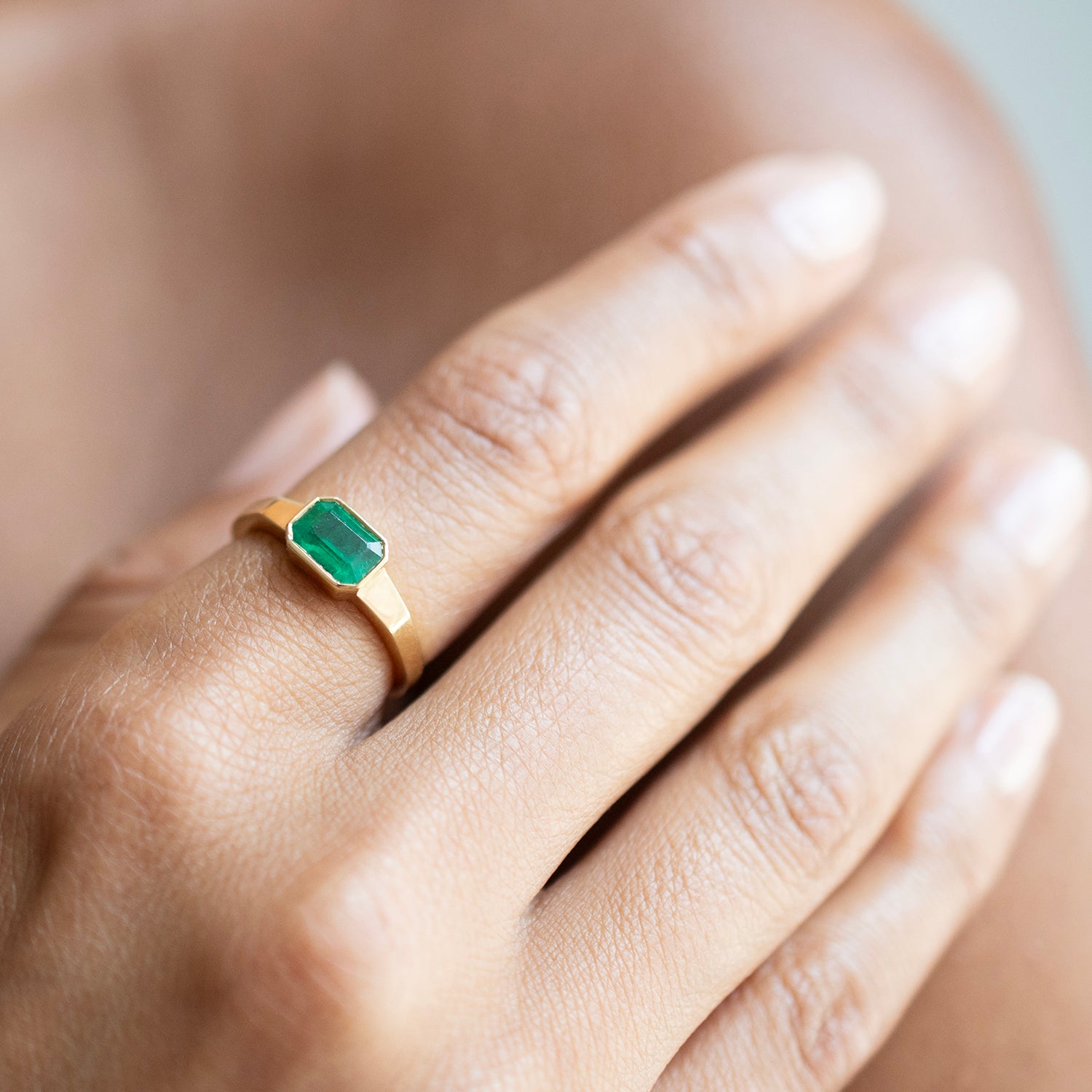 A close-up of a hand wearing a gold ring with a rectangular green emerald-cut gemstone.