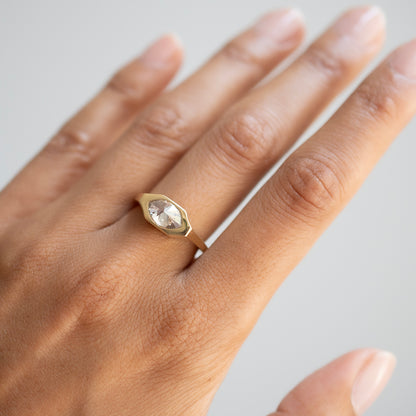 A gold ring with a marquise-cut clear gemstone set in a bezel-style oval shape, shown worn on a person's finger against a soft light gray background.