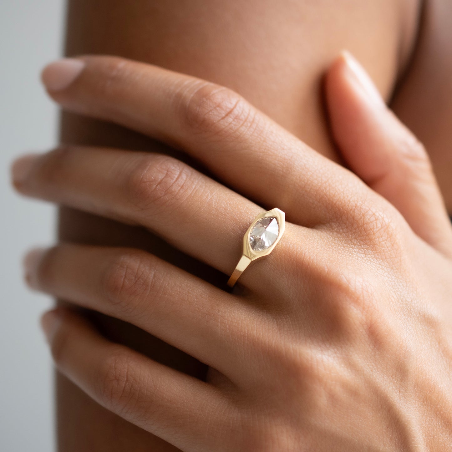 A close-up of a hand wearing a gold ring with a marquise-cut clear gemstone, set against a soft neutral background.