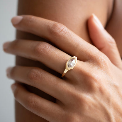 A close-up of a hand wearing a gold ring with a marquise-cut clear gemstone, set against a soft neutral background.
