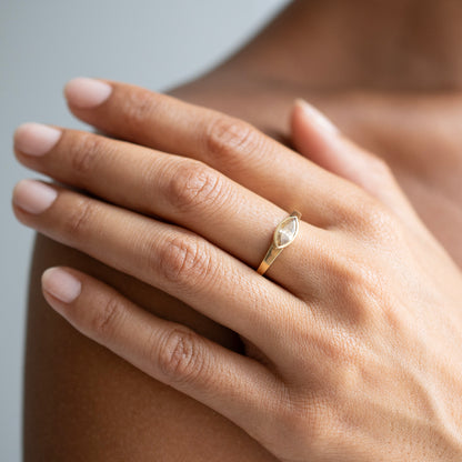 A close-up of a person's hand wearing a delicate gold ring with a marquise-cut clear gemstone, resting on their shoulder.