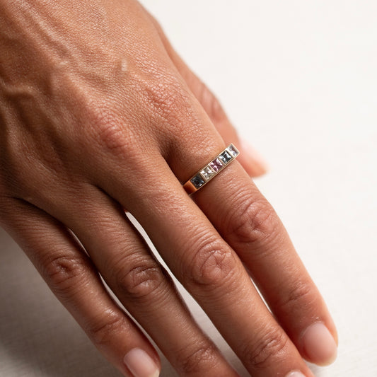 A close-up of a person's hand wearing a slim silver band ring with a row of small clear stones on a plain light background.