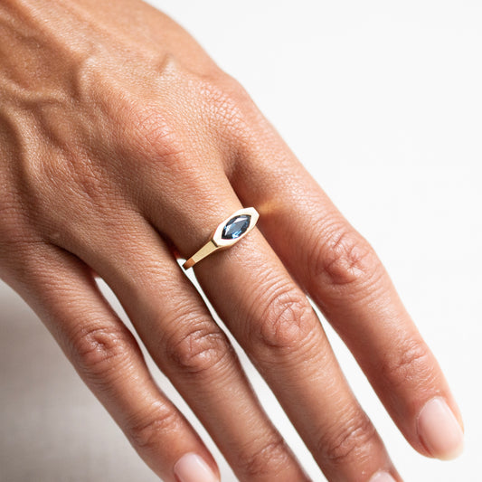 A close-up of a hand wearing a gold ring with a marquise-cut blue gemstone, set against a clean white background.