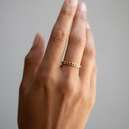 A close-up of a hand wearing a delicate gold band ring with small dark round stones set along the front, against a soft gray background.