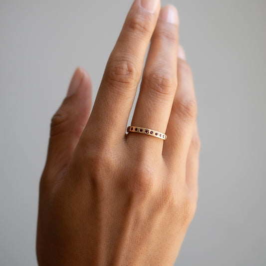 A close-up of a hand wearing a delicate gold band ring with small dark round stones set along the front, against a soft gray background.