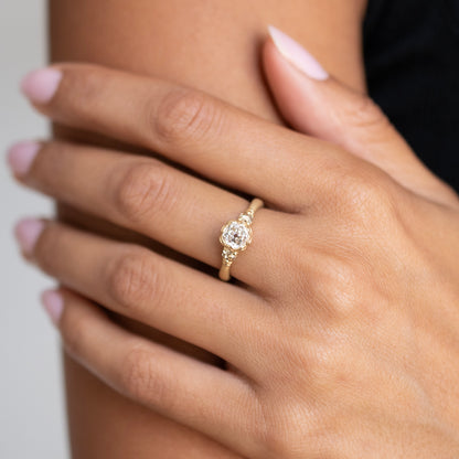 A close-up of a hand wearing a yellow gold ring with a round-cut clear gemstone, set against a soft neutral background.