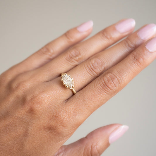 A close-up of a hand wearing a yellow gold ring with a large oval clear gemstone in a decorative bezel setting, against a soft neutral background.