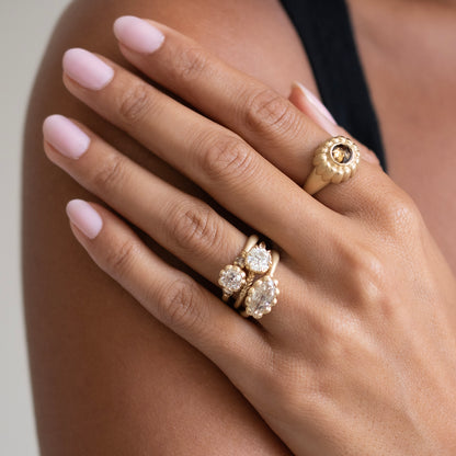 A close-up of a hand wearing two gold rings with clear gemstones, resting on a shoulder. The nails are painted a soft light pink, and a black strap of a top is visible in the background.