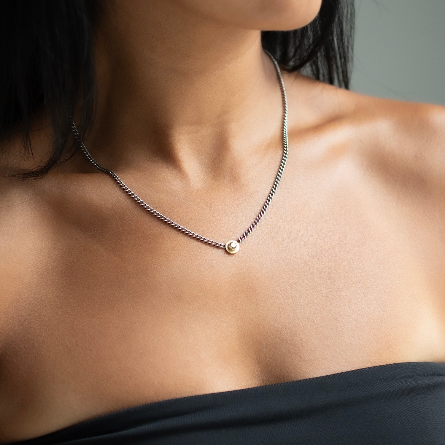 A close-up of a model's neck and upper chest wearing a delicate silver chain necklace with a small gold and diamond pendant.