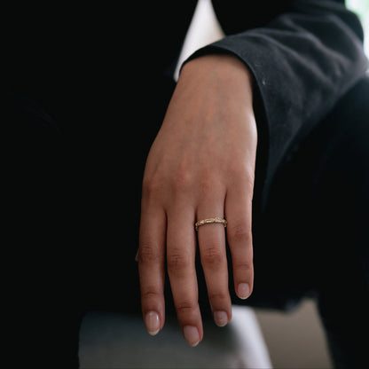 A close-up of a person's hand wearing a delicate gold band ring, dressed in a black blazer against a dark, softly blurred background.