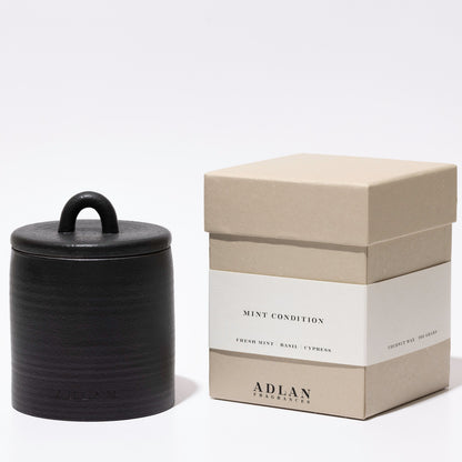 A black ceramic jar candle with a lid and loop handle, displayed next to its beige and white packaging box labeled 'MINT CONDITION' and 'ADLAN COLLECTIONS' on a clean white background.