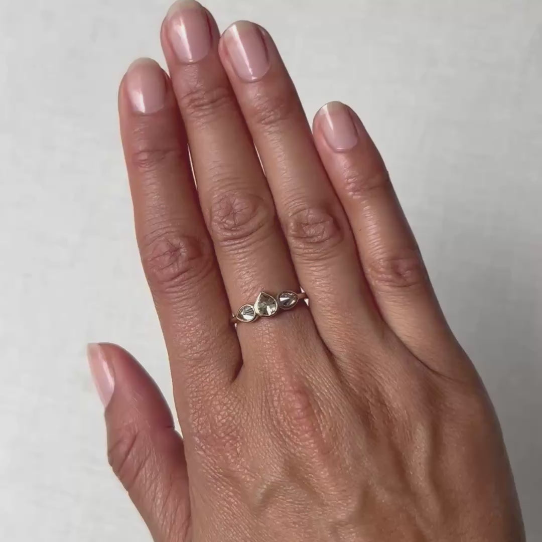 A close-up of a hand wearing a delicate gold ring with three small clear teardrop-cut gemstones, set against a bright white background.