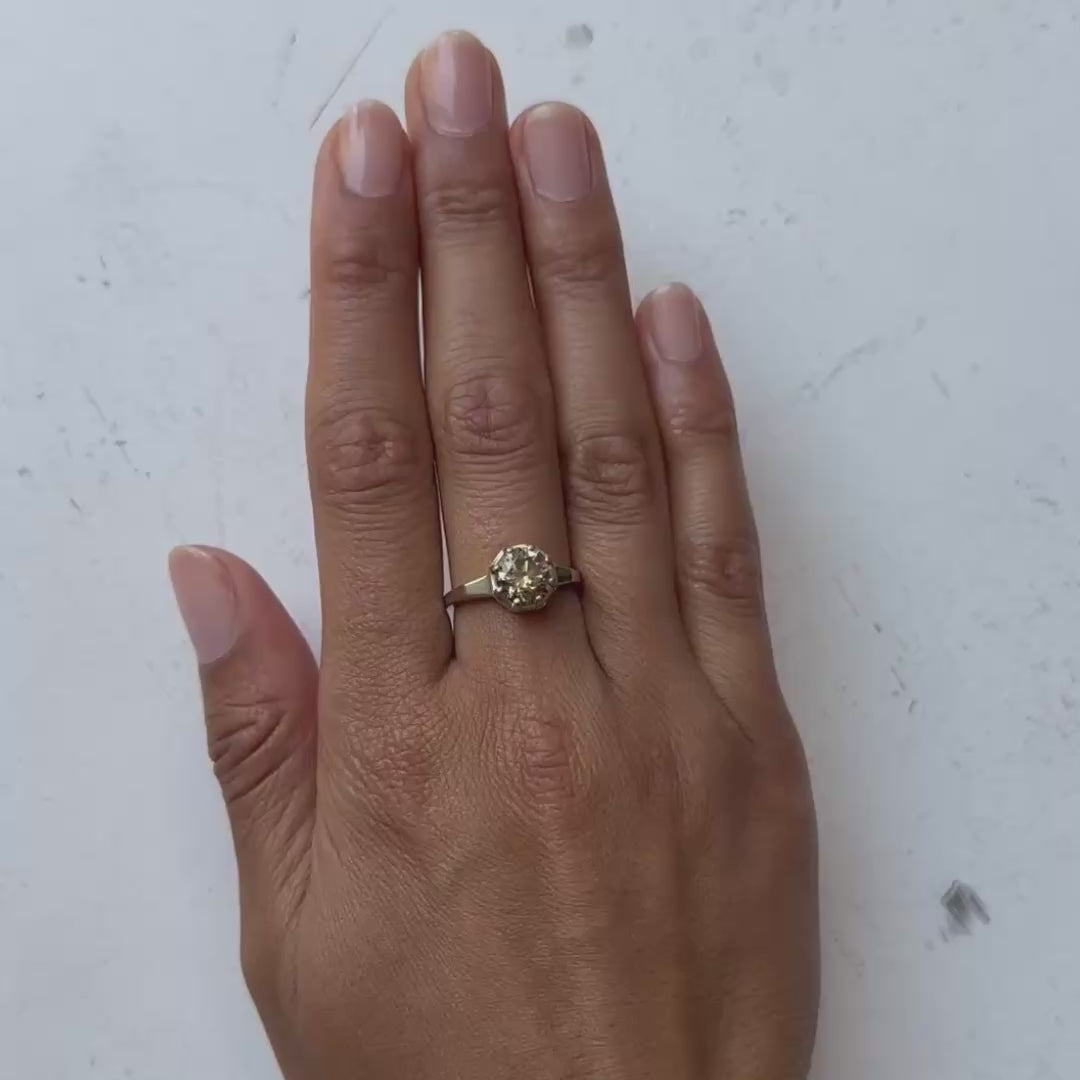 A close-up of a hand wearing a white gold ring with a round clear gemstone, set against a soft light gray background.