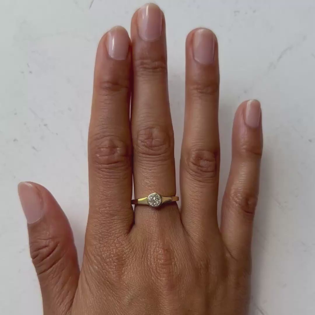 A close-up of a hand wearing a delicate gold ring with a round clear diamond, set against a soft light gray background.