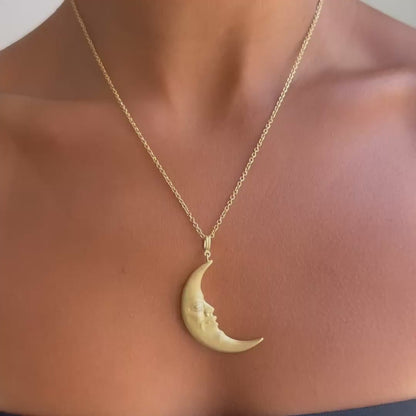 A close-up of a model wearing a delicate gold chain necklace with a crescent moon pendant. The pendant features a small carved face on the side.
